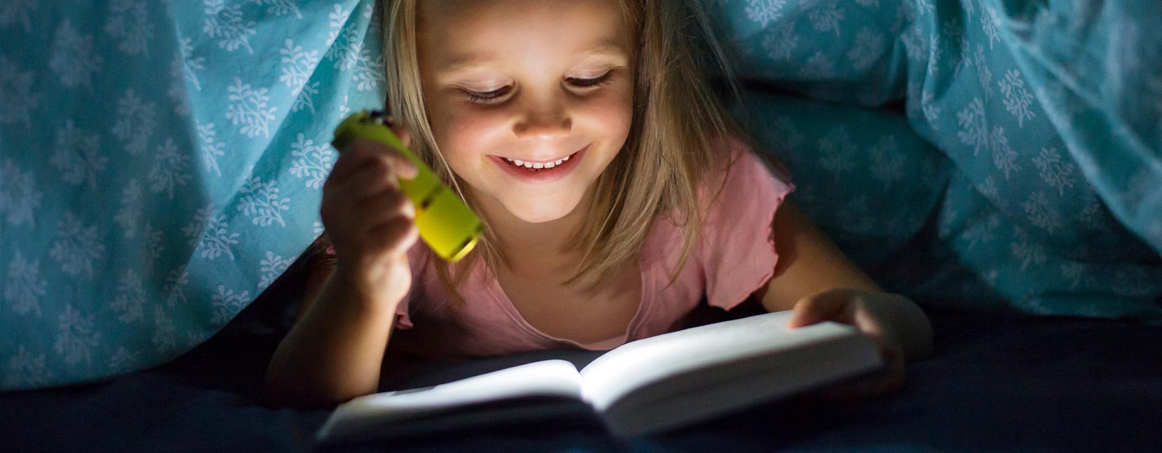 Bambina legge un libro di notte sotto la coperta con una torcia